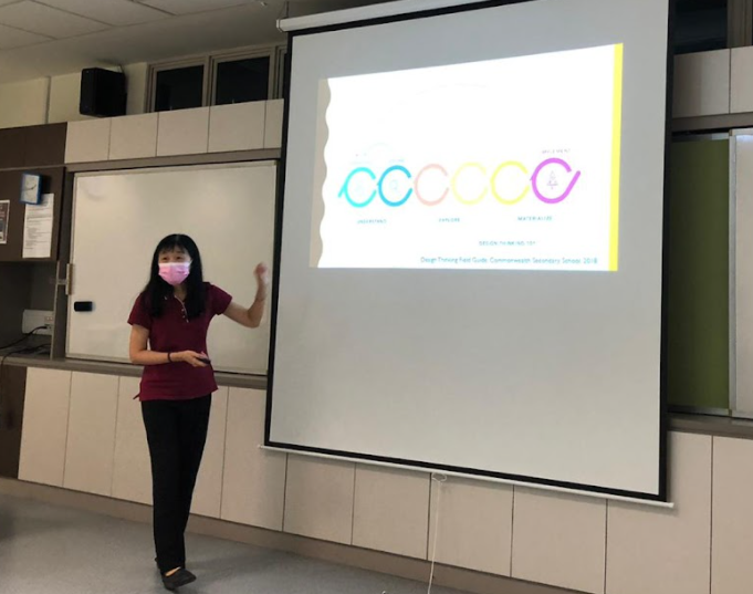 Woman in a red top and pink mask presents beside a projected slide showing colorful circular shapes in a classroom.